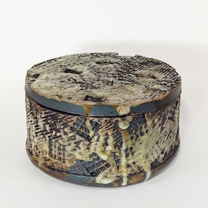 Op de afbeelding: Een ronde keramische doos met een getextureerd deksel en zijkanten. De doos is versierd met een bruine, zwarte en witte glazuur. Het deksel heeft een druppel-effect, waarbij de glazuur langs de zijkanten loopt.