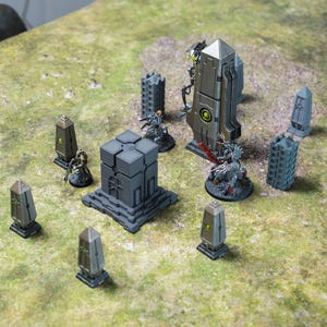 May include: A set of miniature grey and silver buildings, including a tall tower with a yellow accent, a cube-shaped building, and several smaller towers. The buildings are arranged on a green surface.