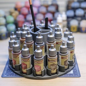 May include: A rotating paint bottle holder with 18 small bottles of paint. The bottles are arranged in a circle and are labelled with different colours and names. The holder is made of grey plastic and has a black base.