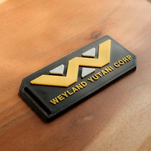 May include: A black plastic badge with a yellow and white logo and the text "Weyland-Yutani Corp."
