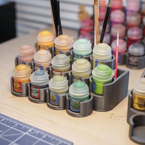 Op de afbeelding: Een 3D-geprinte verfpottenhouder met rijen kleine verfpotten in verschillende kleuren, waaronder groen, oranje en bruin. De houder is gemaakt van grijs plastic en heeft een getextureerd oppervlak.