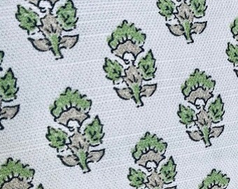 May include: A white fabric featuring a repeating block-printed pattern of small floral motifs. Each motif consists of light green leaves and a central gray stem and bud, arranged in a consistent grid across the textured textile surface.