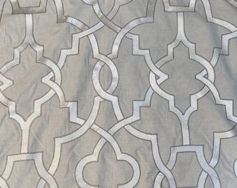 May include: Close-up of a beige fabric with a repeating geometric pattern in silver. The pattern features interlocking shapes and lines, creating a textured, decorative design. The fabric is folded, showing the texture and detail of the embroidery.