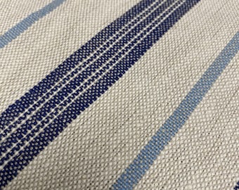 May include: Close-up of a woven fabric with a neutral beige background. Diagonal stripes in varying widths and shades of blue create a pattern. The fabric has a textured appearance, suggesting a natural fiber like linen or cotton.
