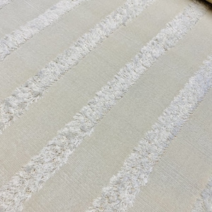 May include: Cream-colored rug with diagonal stripes of textured, white fringe. The rug has a soft, neutral color palette, suitable for various interior design styles. The fringe adds a tactile element to the design.