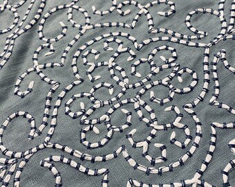 May include: Close-up of a teal fabric with an intricate embroidered design. The embroidery features a repeating pattern of white and navy blue stitched lines, creating a textured, decorative surface. The design is reminiscent of floral or scroll motifs.