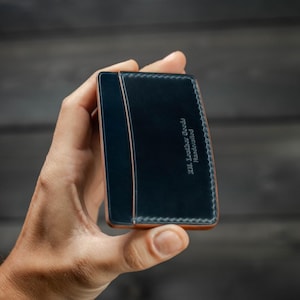 May include: A handcrafted, navy blue leather wallet with a contrasting tan edge and stitching. The wallet has the text "J.M. Leather Goods Handcrafted" embossed on the front. The wallet is held in a hand.