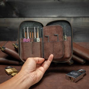 May include: A brown leather lock pick set with various picks and tools, displayed open. The set is in a zippered case with multiple compartments. A hand is holding the case, with a padlock and keys visible nearby.