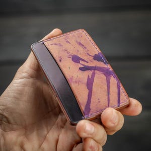 May include: A rectangular leather wallet with a unique design. The wallet features a tan background with purple paint splatter details. The wallet is held in a hand, showcasing its size and design. The wallet appears to be new and unused.