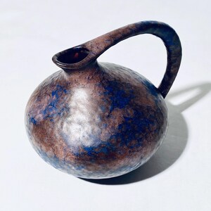 May include: A brown ceramic pitcher with a blue and silver glaze. The pitcher has a rounded body and a curved handle.