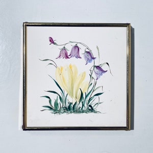 Könnte beinhalten: Ein gerahmtes Kunstwerk mit einer Aquarellmalerei von gelben Krokus und violetten Glockenblumen mit grünen Blättern auf weißem Hintergrund.
