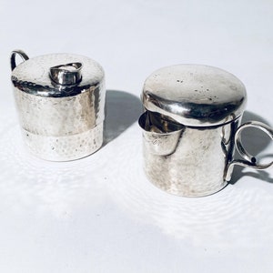 Könnte beinhalten: Ein silbernes Sahne- und Zuckerset mit gehämmerter Textur. Der Milchkännchen hat einen Deckel und einen kleinen Ausguss. Die Zuckerdose hat einen Deckel.