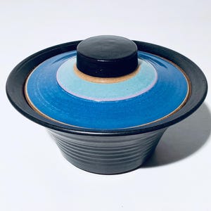 Könnte beinhalten: Eine Keramikschale mit Deckel. Die Schale ist schwarz mit geriffelter Außenseite. Der Deckel ist kreisförmig mit konzentrischen Ringen in Blau, Türkis und Lila. Der Deckel hat einen schwarzen Knopf. Die Schale steht auf einer weißen Oberfläche.