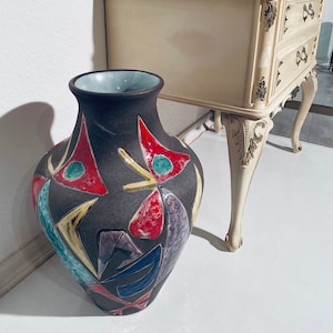 Może przedstawiać: Duży, ciemnobrązowy wazon ceramiczny z geometrycznym wzorem w odcieniach czerwieni, żółci, błękitu i zieleni. Wazon stoi na białym tle obok białego drewnianego stołu z dwoma szufladami.