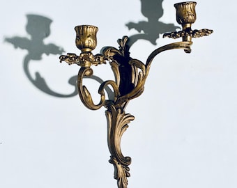 May include: Ornate, antique gold-colored wall sconce with two candle holders. The sconce features a detailed, scrolling design with floral accents. The sconce is mounted on a white wall, casting a shadow.