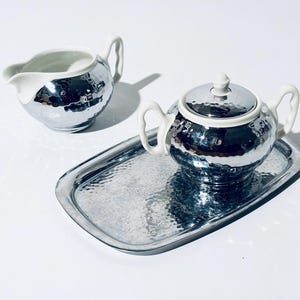 May include: A silver tea set with a hammered texture, including a teapot with a lid, a creamer, and a rectangular tray. The teapot and creamer have white accents and handles. The set is designed for serving tea.