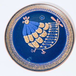May include: A round ceramic plate with a blue background and a stylized chicken design. The chicken is depicted in shades of blue, yellow, and orange. The plate has a brown rim and a decorative border. The plate is a decorative item.