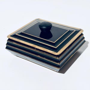 Könnte beinhalten: Eine quadratische, schwarze Box mit goldenem Rand und einem kleinen, runden, schwarzen Knopf auf dem Deckel. Die Box hat ein mehrschichtiges Design mit goldenen Akzenten. Die Box wird wahrscheinlich zur Aufbewahrung kleiner Gegenstände verwendet.