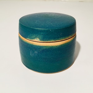 Könnte beinhalten: Eine tealfarbene Keramikdose mit Deckel. Die Dose hat einen hellbraunen Rand und eine subtile Glasurvariation.