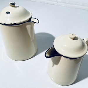 Könnte beinhalten: Zwei weiße Emaille-Kannen mit schwarzem Rand. Die größere Kanne hat einen Deckel und einen Ausguss. Die kleinere Kanne hat einen Deckel und einen Griff.