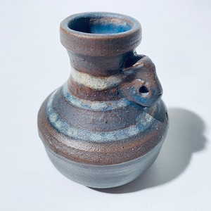 Könnte beinhalten: Eine kleine, braune Keramikvase mit blauen und weißen Streifen. Die Vase hat einen einzigartigen Griff in Form eines kleinen Tierkopfes.