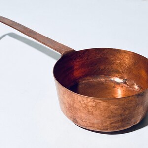 Könnte beinhalten: Eine antike Kupfer Schöpfkelle mit einem langen, gebogenen Griff. Die Schöpfkelle ist angelaufen und hat ein rustikales Aussehen.