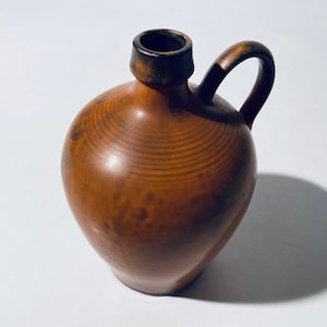 Może przedstawiać: Brązowy dzbanek ceramiczny o zaokrąglonym korpusie i uchwycie. Dzbanek ma fakturowaną powierzchnię i ciemnobrązową krawędź.