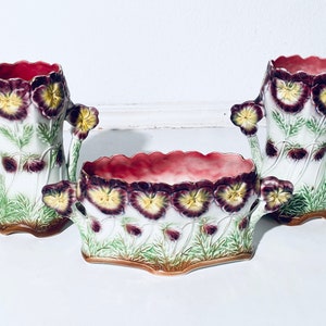 May include: A set of three vintage ceramic vases with a floral design. The vases are decorated with hand-painted pansies in shades of purple, yellow, and green. The vases are in excellent condition and would make a beautiful addition to any home.