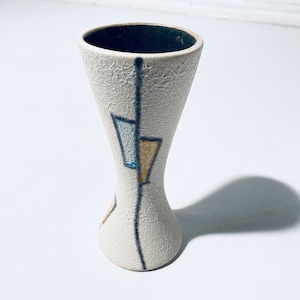 Könnte beinhalten: Eine weiße Keramikvase mit einem geometrischen Design in Blau, Gelb und Schwarz. Die Vase hat eine strukturierte Oberfläche und eine schmale Öffnung.