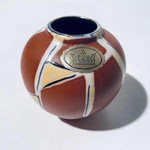 Könnte beinhalten: Eine kleine, runde, terracottafarbene Vase mit einem geometrischen Muster in Weiß, Gelb und Schwarz. Die Vase hat ein goldenes Etikett mit einer Krone und dem Wort "Bongo" darauf.