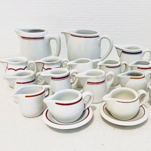 Könnte beinhalten: Eine Sammlung weißer Keramik-Sahnekännchen und kleiner Servierschalen. Die Kännchen variieren in Größe und Form, mit einem roten Streifen um den Rand. Einige haben passende Untertassen. Die Sammlung ist auf einer weißen Oberfläche angeordnet.