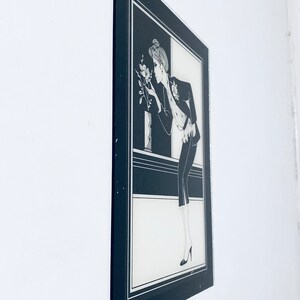 May include: Black and white framed illustration of a woman in a suit, looking at a flower. The woman is wearing a hat and has her hair pulled back. The background is a black and white geometric pattern.