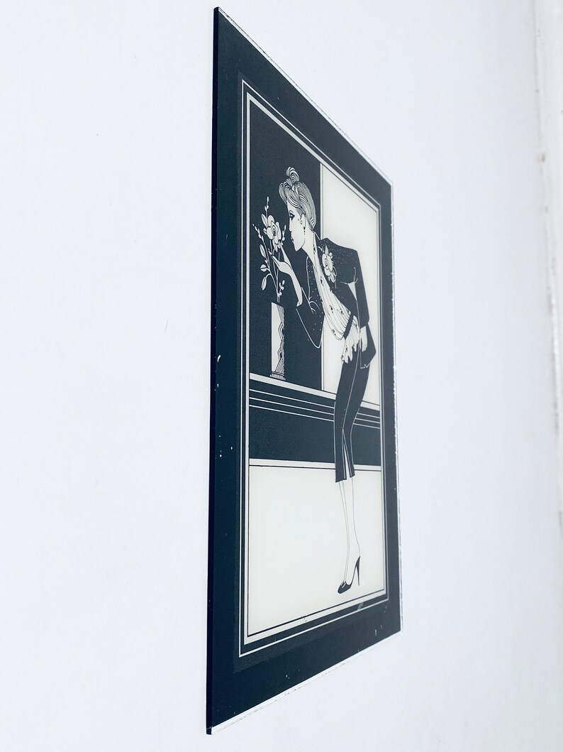 May include: Black and white framed illustration of a woman in a suit, looking at a flower. The woman is wearing a hat and has her hair pulled back. The background is a black and white geometric pattern.