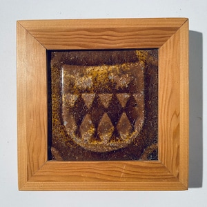 Könnte beinhalten: Eine gerahmte Keramikfliese mit brauner und gelber Glasur. Die Fliese zeigt ein erhabenes Design eines Schildes mit drei stilisierten Dreiecken.