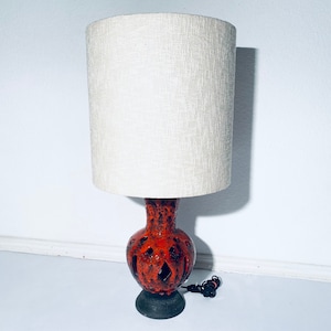 Puede incluir: Una lámpara de mesa de cerámica con un esmalte marrón y naranja. La lámpara tiene una base redonda con un diseño recortado y una pantalla de tela blanca.