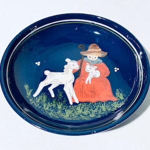 Op de afbeelding: Een blauw keramiek bord met een witte rand en een geschilderd tafereel van een meisje in een rode jurk dat een lam vasthoudt. Het meisje draagt een hoed en houdt een herdersstaf vast. Er zijn twee lammeren in de scène, een in de armen van het meisje en een dat naast haar staat.