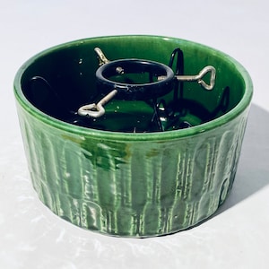 Könnte beinhalten: Ein grüner Keramik-Weihnachtsbaumständer mit einem Metall-Baumhalter. Der Ständer hat ein strukturiertes, vertikales Muster.