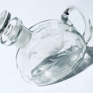 Puede incluir: Una decantador de vidrio transparente con un diseño grabado de flores y un tapón de vidrio. La decantador tiene un asa y tiene forma de corazón.