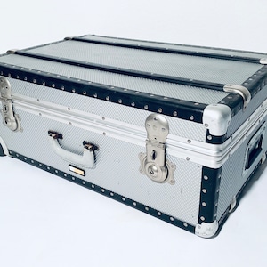 Könnte beinhalten: Silberner Rollkoffer mit schwarzem Besatz und Metallbeschlägen. Der Koffer hat eine strukturierte Oberfläche, einen Griff und Eckschützer. Er verfügt über einen Schnappverschluss und Rollen.