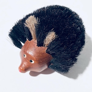 Könnte beinhalten: Holz-Igel-Figur mit schwarzen Borsten und beigen Akzenten. Der Igel hat einen braunen Körper, kleine weiße Augen und eine rosa Nase. Dieser Dekorationsartikel ist wahrscheinlich eine Bürste oder eine Sammlerfigur.