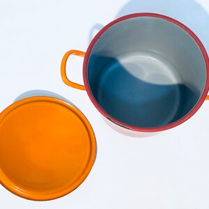 Pu&ograve; includere: Una pentola di smalto arancione con un bordo rosso e un coperchio arancione abbinato. La pentola ha due manici ed &egrave; vuota.