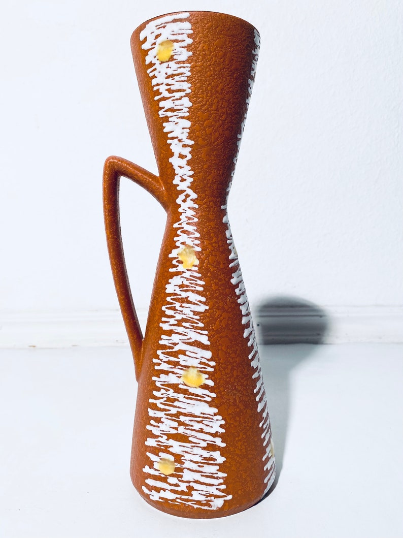 Puede incluir: Un jarr&oacute;n de cer&aacute;mica marr&oacute;n con un patr&oacute;n geom&eacute;trico blanco y amarillo. El jarr&oacute;n tiene una abertura acampanada y un asa.