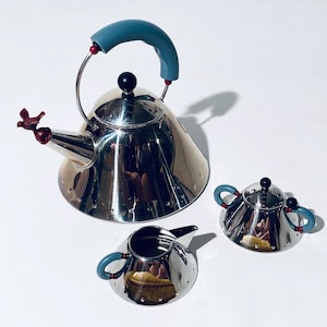 Könnte beinhalten: Ein Edelstahl-Teeservice mit Teekanne, Sahnekännchen und Zuckerdose. Die Teekanne hat einen blauen Griff und einen vogelförmigen Ausguss. Sahnekännchen und Zuckerdose haben blaue Griffe. Alle Teile haben eine glänzende, reflektierende Oberfläche.