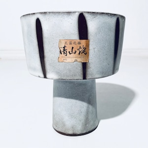 Op de afbeelding: Een keramische voetkom met een lichtgrijze glazuur en verticale donkergrijze strepen. Een klein rechthoekig label met Japanse tekst is op de kom bevestigd. De kom staat op een korte, cilindrische voet.