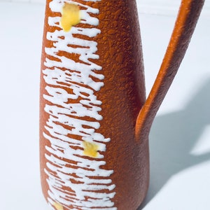 Puede incluir: Una jarra de cer&aacute;mica marr&oacute;n con un esmalte blanco y amarillo. La jarra tiene una superficie texturizada y un asa.