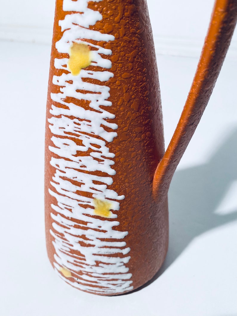 Puede incluir: Una jarra de cer&aacute;mica marr&oacute;n con un esmalte blanco y amarillo. La jarra tiene una superficie texturizada y un asa.