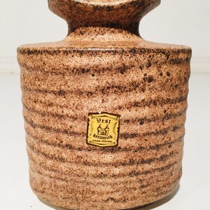 Może przedstawiać: Brązowy ceramiczny wazon z fakturowaną powierzchnią i etykietą z napisem "Vest keramiek Gouda-Holland".
