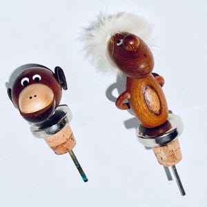 Könnte beinhalten: Zwei skurrile Weinflaschenverschlüsse aus Holz. Einer ist ein Affengesicht, der andere eine Figur mit einem flauschigen weißen Kopf. Beide haben Korkbasen, Metallringe und Metallschnäbel. Der Affenverschluss hat ein braunes Gesicht und Ohren.