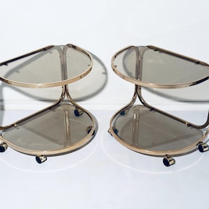 May include: Two gold-toned, two-tiered serving carts with smoked glass shelves. Each cart has a curved, geometric design and sits on small black wheels. The carts are designed for serving drinks or displaying items.