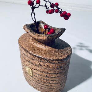 Może przedstawiać: Brązowy i czarny, pstrokaty ceramiczny wazon o nietypowym kształcie z złotą etykietą. Wazon jest wypełniony gałązką czerwonych jag&oacute;d.